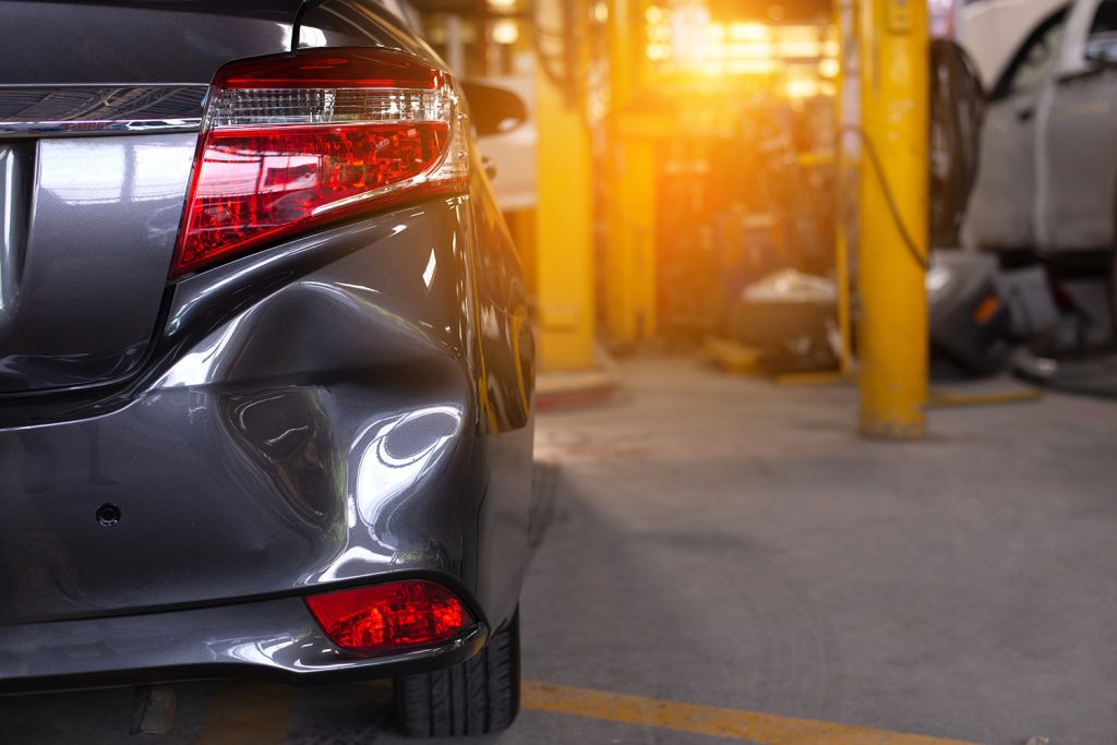 A close-up of a black car’s back right, showing a large dent in the bumper, inside of a body shop.