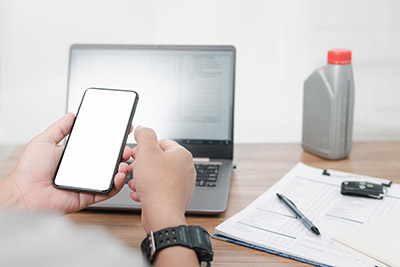 A person holding a smartphone with a laptop, car keys, pen, paper, and oil carton in the background.